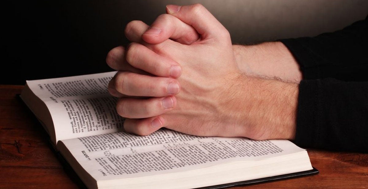 Hands folded in prayer over a Holy bible on wooden table on grey background